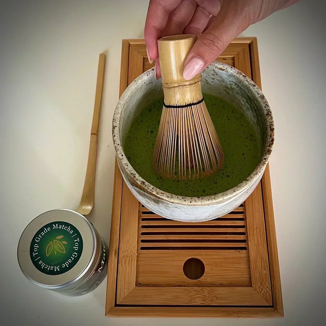 Hand met Japanse bamboe matcha klopper roerend in ecru matcha kom met matcha op serveerblaadje, een bamboelepel en een blikje 'Ceremonial Matcha' van dThe Matcha Man.