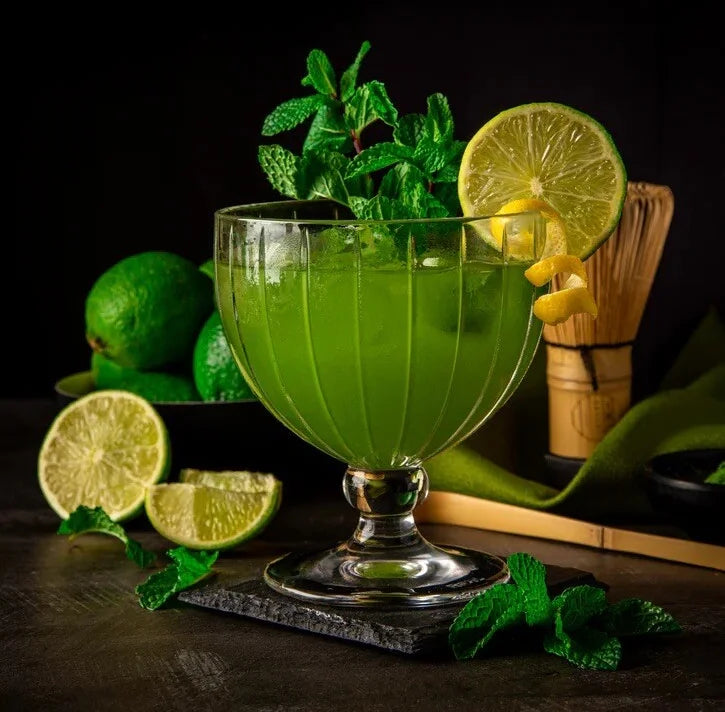 Een groene cocktail met matcha gedecoreerd met een schijfje citroen en muntblaadjes, daarnaast een bamboe matcha klopper, een  bamboelepel en citrusvruchten.