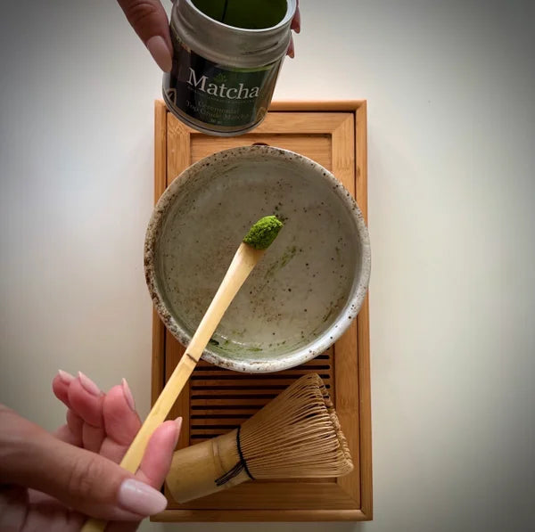 Hand met bamboelepel en matcha boven een matcha kom en serveerblaadje en een blikje matcha van The Matcha Man.