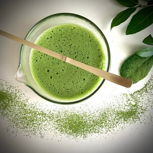 Een met matcha gevulde glazen matcha  kom  met tuit en een bamboelepel.