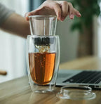 Dubbelwandig theeglas gevuld met thee en hand met glazen theefilter met losse thee.
