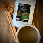 Zakje 'Premium Hojicha latte' van The Matcha Man uit Japan, een witte matcha kom gevuld met hojicha en een bamboe matcha klopper op een serveerblaadje.