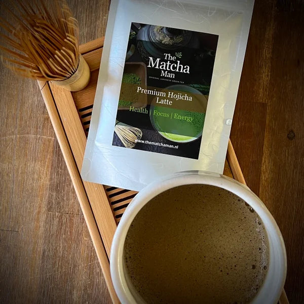 Zakje 'Premium Hojicha latte' van The Matcha Man uit Japan, een witte matcha kom gevuld met hojicha en een bamboe matcha klopper op een serveerblaadje.