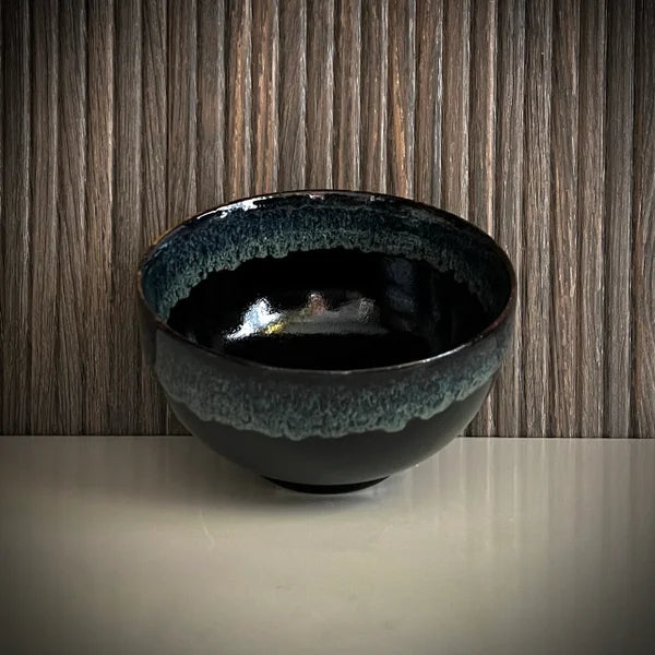 Voorkant van een geglazuurde zwarte matcha kom van keramiek met groen-blauwe rand.