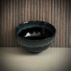 Voorkant van een geglazuurde zwarte matcha kom van keramiek met groen-blauwe rand.