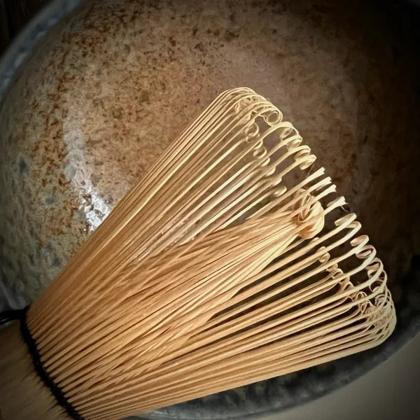 Gedeelte van een bamboe matcha klopper met beige matcha kom met zachtblauwe rand op de achtergrond.