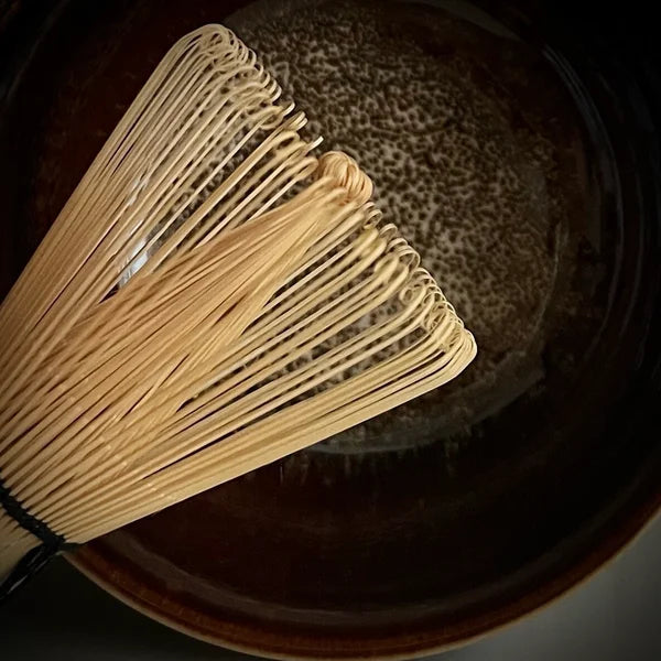 Gedeelte van een bamboe matcha klopper met een bordeaux matcha kom op de achtergrond.