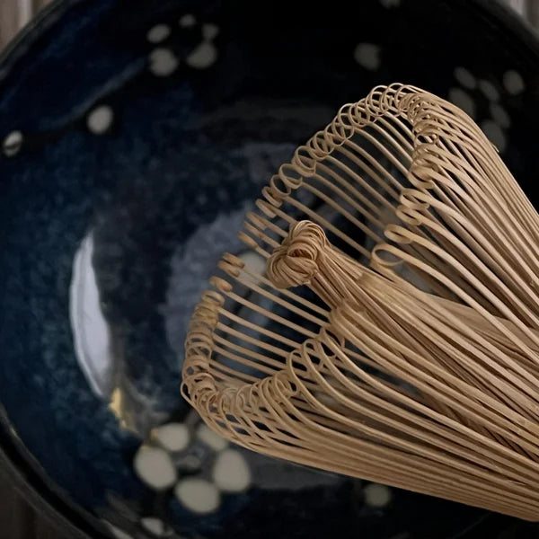 Gedeelte van een bamboe matcha klopper met een donkerblauwe matcha kom met bloesemtakken op de achtergrond.