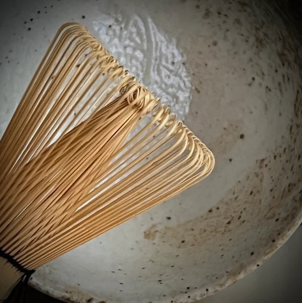 Gedeelte van een bamboe matcha klopper met ecru matcha kom op de achtergrond.