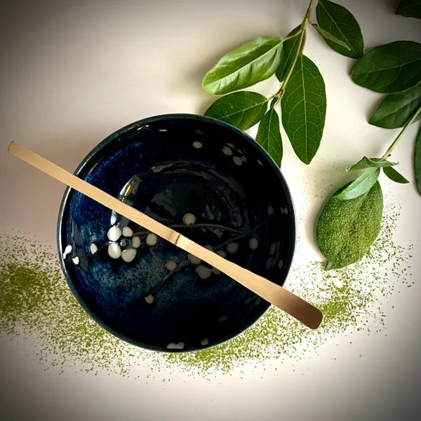 Bovenaanzicht van een donkerblauwe matcha kom van keramiek met bloesemtakken en een bamboelepel.