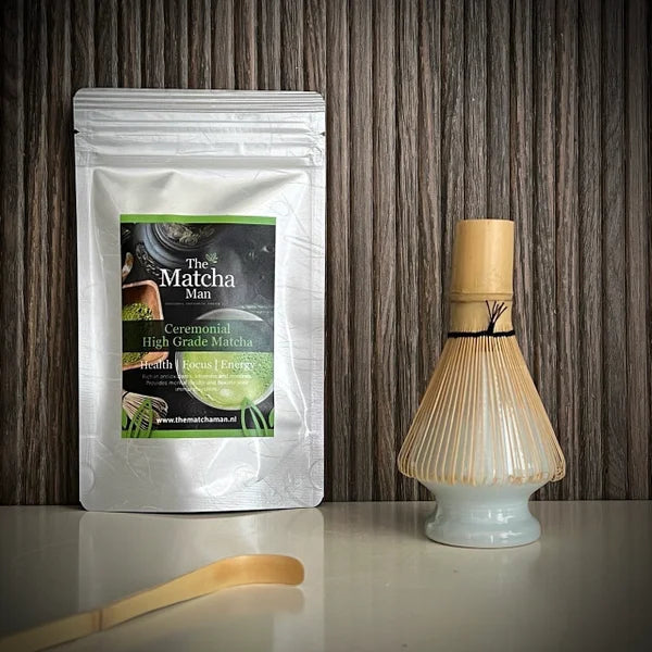 Hersluitbaar zakje Ceremonial High Grade Matcha, een bamboe matchaklopper op een lichtblauwe whiskhouder en neen bamboelepel op de voorgrond.