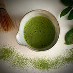 Een ecru chawan met schenktuit gevuld met matcha en een gedeelte van een bamboe matcha klopper.