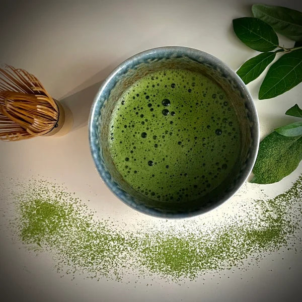 Bovenaanzicht van een beige matcha kom van keramiek met blauw-groene rand gevuld met matcha en een bamboe matcha klopper.