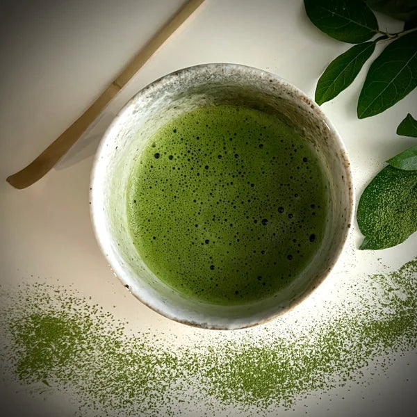 Bovenaanzicht van een ecru matcha kom van keramiek met groen-bruine tinten gevuld met matcha en een bamboe matcha klopper.