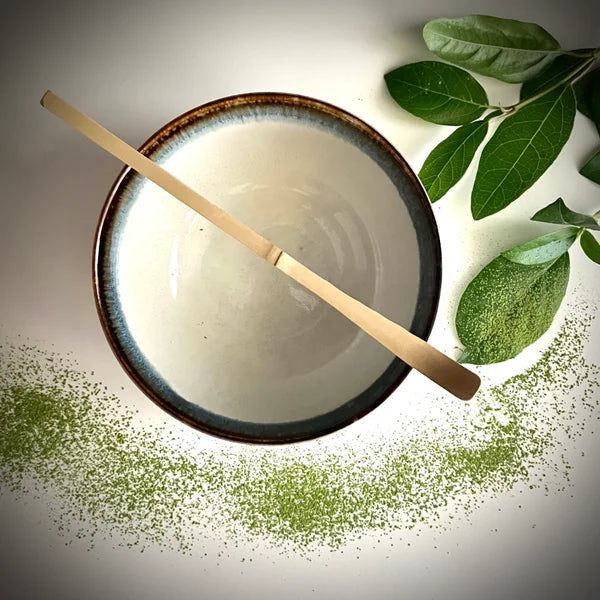 Lcihtblauwe matcha kom met bruine rand en een bamboelepel.
