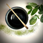 Bovenaanzicht van een geglazuurde zwarte matcha kom van keramiek met groen-blauwe rand en bamboelepel.