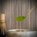 Een bamboe matcha klopper  op whiskhouder en een bamboelepel met matcha boven een gedeelte van een match kom.