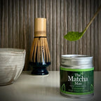Zilverkleurig potje 'Ceremonial High Grade Matcha' van The Matcha Man (30 gram), een gedeelte van een matcha kom met tuit, een bamboe matcha klopper op een whiskhouder en een bamboelepel met matcha.