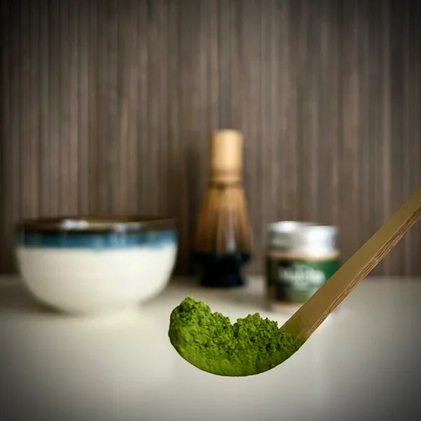 Bamboelepel met losse matcha er op en op achtergrond een matcha kom, een bamboe matcha klopper en potje matcha.