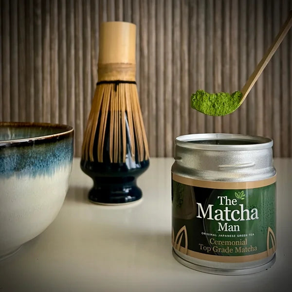 Zilverkleurig potje 'Ceremonial Top Grade Matcha' van The Matcha Man (30 gram), een gedeelte van een lichtblauwe matcha kom, een whisk op een whiskhouder en bamboelepel met matcha.