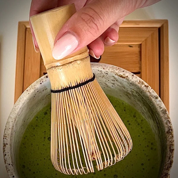 Hand met Japanse bamboe matcha klopper roerend in ecru matcha kom met matcha op serveerblaadje.