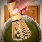 Hand met Japanse bamboe matcha klopper roerend in ecru matcha kom met matcha op serveerblaadje.
