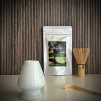 Hersluitbaar zakje Ceremonial High Grade Matcha, een bamboe matchaklopper, een bamboelepel een lichtblauwe whiskhouder op voorgrond.