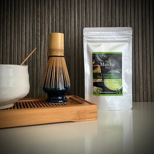 Hersluitbaar zakje 'Ceremonial High Grade Matcha' van The Matcha Man (25 gram), een bamboe matcha klopper op zwarte whiskhouder en gedeelte van een matcha kom op een serveerblaadje.