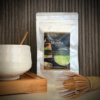 Hersluitbaar zakje 'Ceremonial Top Grade Matcha' van The Matcha Man (25 gram) , een bamboe bamboe matcha klopper en een witte matcha kom op een serveerblaaadje.